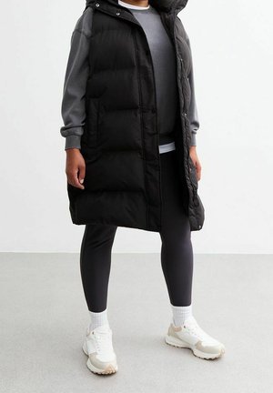 Personne portant une longue doudoune noire sans manches par-dessus un sweat gris, des leggings noirs, des chaussettes blanches et des baskets blanches, debout sur un sol gris.