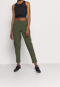 Black sleeveless top, green tapered pants, and black athletic shoes. Cotton material with a relaxed fit. Notable logo on the top.