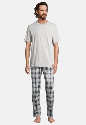 Uomo con la barba in piedi a piedi nudi che indossa una camicia a maniche corte grigio chiaro e pantaloni del pigiama a quadri bianchi e neri su uno sfondo semplice.