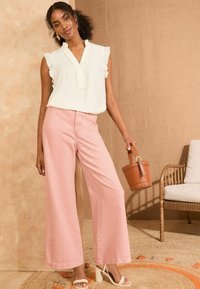 Blusa bianca senza maniche con orli arruffati, abbinata a pantaloni rosa a gamba larga. La modella tiene una borsa a secchiello marrone. Sfondo naturale.