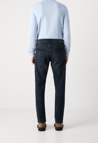 Hombre con jeans azul oscuro, camisa de manga larga azul clara y zapatos marrones de pie de espaldas sobre un fondo blanco liso.