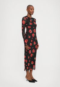 Svart blomsterkjole med lange gjennomsiktige ermer, med røde og grønne blomstermønstre. Slim fit, knelangs, og gjennomsiktige detaljer i kanten.