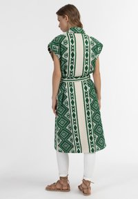 Zaļš un krēms ar rakstu midi kleita ar īsām piedurknēm, jostu virs vidukļa un ģeometrisku dizainu, kombinēta ar baltām biksēm un sandālēm.