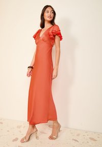 Sarkana maxi kleita ar volānveida piedurknēm, dziļu V-veida izgriezumu un gludu tekstūru. Valkāta kopā ar metāliskām sandaletēm ar siksniņām, uz gaiša fona.