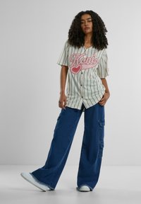 Stripete baseballtrøye i kremfarge med rosa bokstaver, paret med vide blå cargo-bukser som har lommer og frynsete kanter. Hvite sneakers.