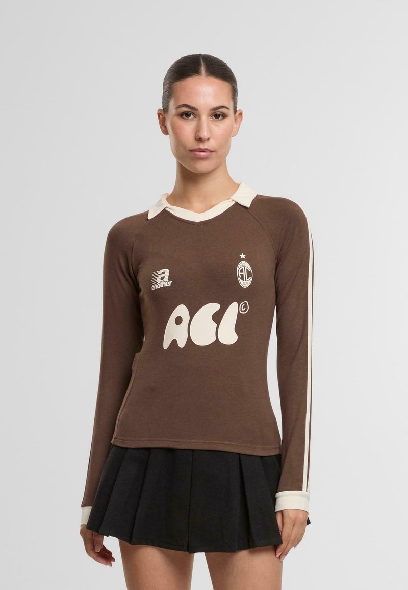 Ruskea pitkähihaisella paidalla, jossa on valkoinen kaulus, suuret valkoiset tekstit ja logo. Yhdessä pleatattujen mustien hameiden kanssa.