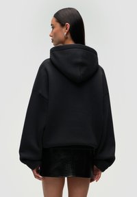 Czarna, oversize bluza z kapturem, z długimi rękawami i gładkiego, strukturalnego materiału. Noszona z błyszcząca czarną mini spódnicą.