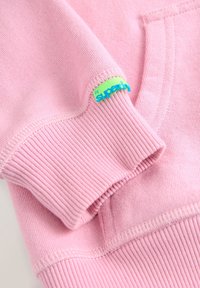 Tela de sudadera rosa claro con puños acanalados; presenta una pequeña etiqueta con un logotipo verde y azul. Textura suave y diseño casual.