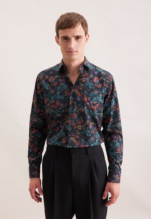 Bluse mit Blumenmuster in Dunkelblau, mit mehrfarbigen Blumen, langen Ärmeln und klassischem Kragen. Kombiniert mit schwarzen Hosen.