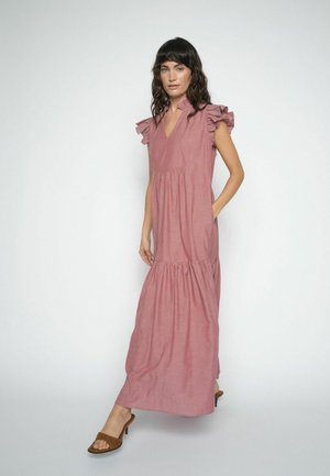 Žena stojaca v bezrukávových maxi šatách ružovej farby so zložitej štruktúre na ramenách a hnedých sandáloch s otvorenou špičkou pred jednoduchým bielym pozadím.