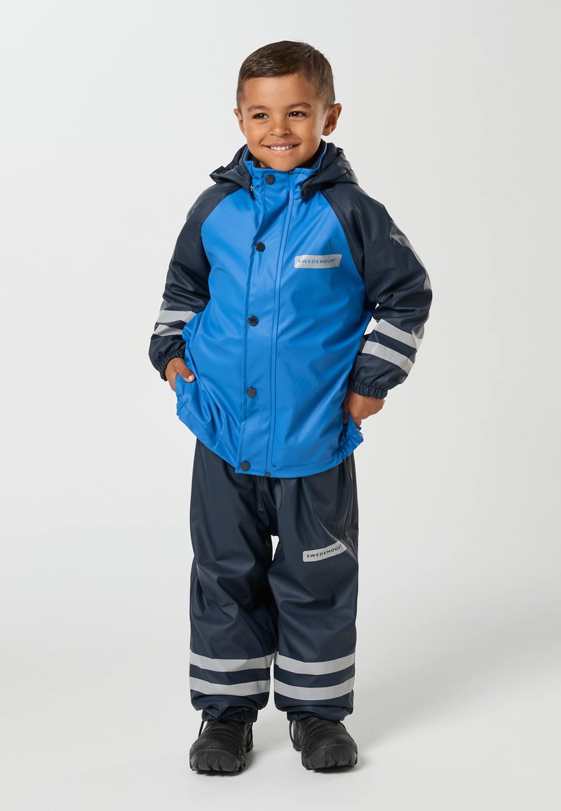 Ung dreng smiler, iført blå og mørkeblå vandtæt jakke og bukser med reflekstriber og sorte støvler.