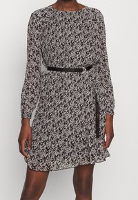 Zwart-witte jurk met bloemenpatroon, lange mouwen, geplooide details, een gecinchte taille en een zwarte riem die de taille accentueert.