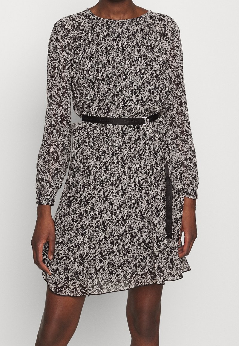 Zwart-witte jurk met bloemenpatroon, lange mouwen, geplooide details, een gecinchte taille en een zwarte riem die de taille accentueert.