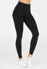 Hochtaillierte schwarze Leggings aus elastischem Stoff, mit glatter Textur und figurbetontem Design, kombiniert mit weißen Sportschuhen.