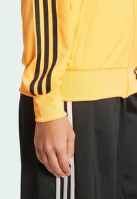 Gelbe Zip-Jacke mit schwarzen Streifen an den Ärmeln, kombiniert mit schwarzen Hosen mit weißen Seitenstreifen. Weicher, strukturierter Stoff.