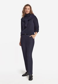 Marineblauer Rollkragenpullover mit drapierter Gestaltung, kombiniert mit einer passenden, enganliegenden Hose, abgerundet durch glänzende schwarze Schuhe.