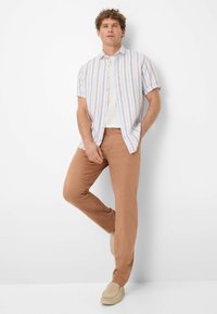 Kurzärmliges, knöpfbares Hemd mit hellblauen und beigen Streifen, getragen über einem weißen T-Shirt. Kombiniert mit hellbraunen Hosen und beigen Slippern.