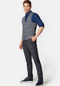 Kamizelka w szary wzór hounds-tooth o dopasowanym kroju, zestawiona z ciemnymi spodniami w kratę i granatowymi sneakersami. Gładka tekstura i wyraźne linie.