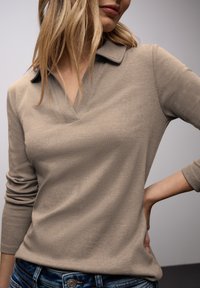 Femme aux cheveux blonds portant un polo beige côtelé à manches longues et un jean bleu, main posée sur la hanche, fond neutre.