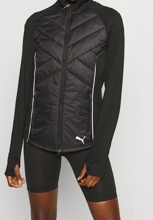 Sort lynlåstrøje med polstret frontpanel og hvidt Puma-logo, båret med sorte cykelshorts på en person mod en ensfarvet baggrund.