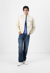 Kremfarget fleecejakke med høy krage, glidelås og logoakkcent. Bæres over en blå hettegenser, sammen med mørke jeans og hvite sko.