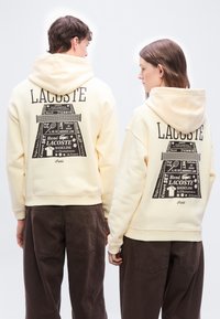 Δύο άτομα φοράνε κρεμ φούτερ με κουκούλα με μεγάλα μαύρα γραφικά τένις της Lacoste στην πλάτη, σε συνδυασμό με σκούρα καφέ παντελόνια.