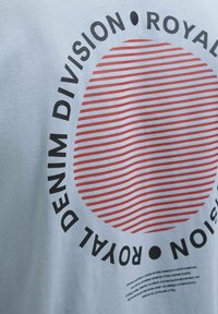 Šviesiai mėlynas trumpomis rankovėmis marškinėliai su raudonų dryžuotų apskritimų dizainu ir ryškiu juodu tekstu "ROYAL DENIM DIVISION." Medvilnės audinys.
