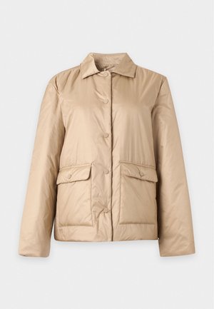 Femme debout, les mains dans les poches, portant une veste beige, un col roulé court rayé et un pantalon cargo noir à jambes larges sur fond blanc.