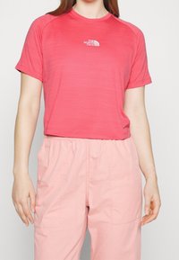 Krátký rukáv, zkrácené tričko v živé korálové barvě s texturovaným povrchem. Na hrudi je malé bílé logo. Kombinováno se světle růžovými kalhotami.