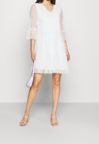 Robe blanche avec manches transparentes et ourlet à volants. Encolure en V, design superposé, associée à un sac violet clair et des sandales à talons beiges.