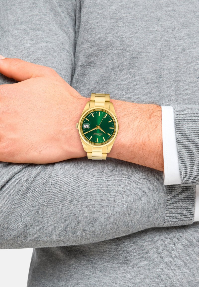 Goldene Armbanduhr mit grünem Zifferblatt, ausgestattet mit einem Datumsdisplay, metallichem Band und minimalistischen Stundenmarkierungen, getragen an einer Hand gegen einen grauen Pullover.
