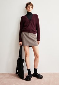 Frau in einem burgunderfarbenen Cardigan über einem schwarzen Rollkragenpullover, grauem Rock, schwarzen Socken und schwarzen Mokkasins, die drinnen steht und eine schwarze Tasche hält.