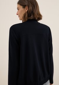 Veste noire à manches longues, à coupe décontractée, avec un ourlet côtelé et une texture de tissu lisse. Comprend un détail de conception avec yoke au dos pour plus de structure.