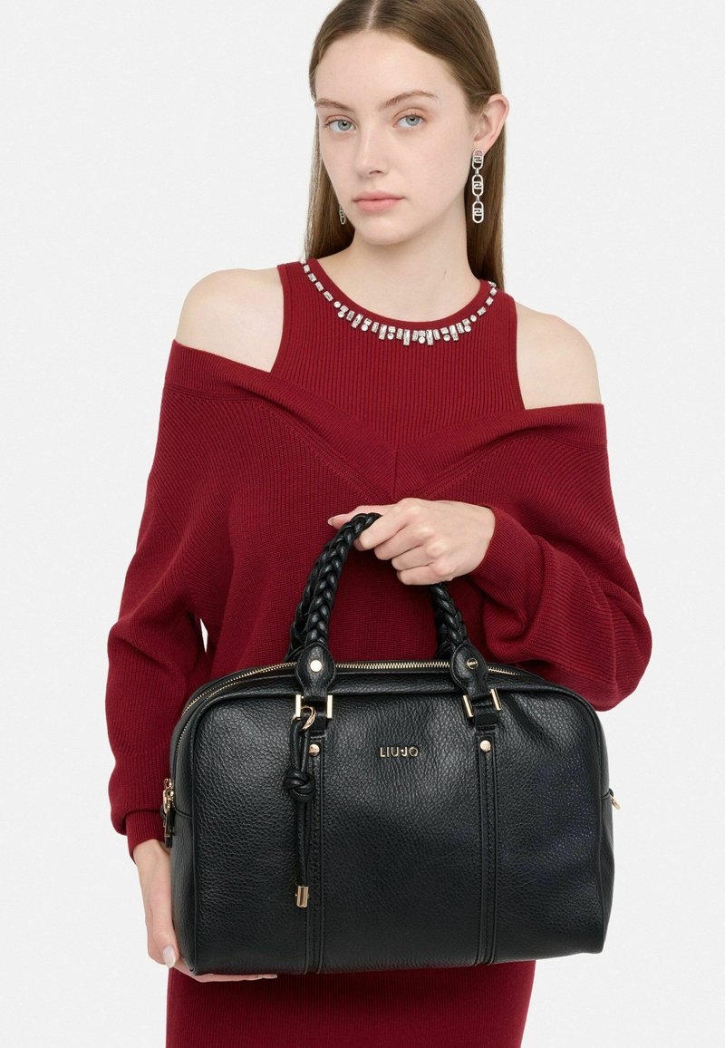 Sac à main en cuir noir avec des anses en cuir tressé et des accents en métal doré. Présente une surface texturée et le logo de la marque sur le devant.