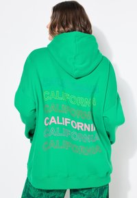 Vihreä huppari pehmeästä kankaasta, jossa on suuri huppu ja monivärinen "CALIFORNIA" -teksti painettuna selässä eri kokoisina.