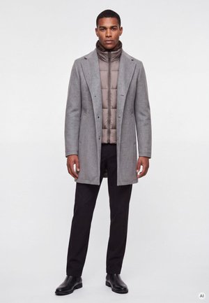 Mann trägt einen grauen Mantel über einer beigen Daunenjacke, schwarze Hose und schwarze Lederstiefel, steht vor einem einfachen weißen Hintergrund.