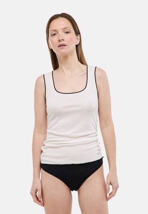 Femme portant un débardeur blanc côtelé sans manches avec un bord noir et un bas de bikini noir, debout devant un fond uni.