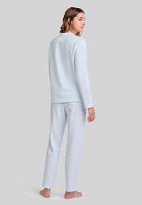 Mujer de pie descalza, vistiendo una blusa de manga larga azul clara y pantalones a juego, de perfil frente a un fondo gris liso.