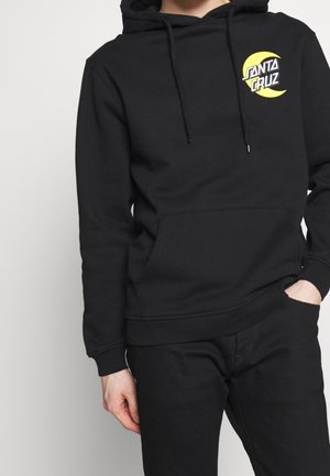 Person iført en sort hættetrøje med en gul halvmåne og "Santa Cruz"-logo på brystet, kombineret med sorte bukser.