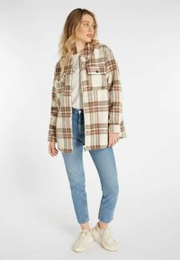 Overmaatse geruite jas in crème, bruin en beige. Beschikt over twee voorzakken, knoopdetails en een relaxte pasvorm. Gepaarde met blauwe jeans.