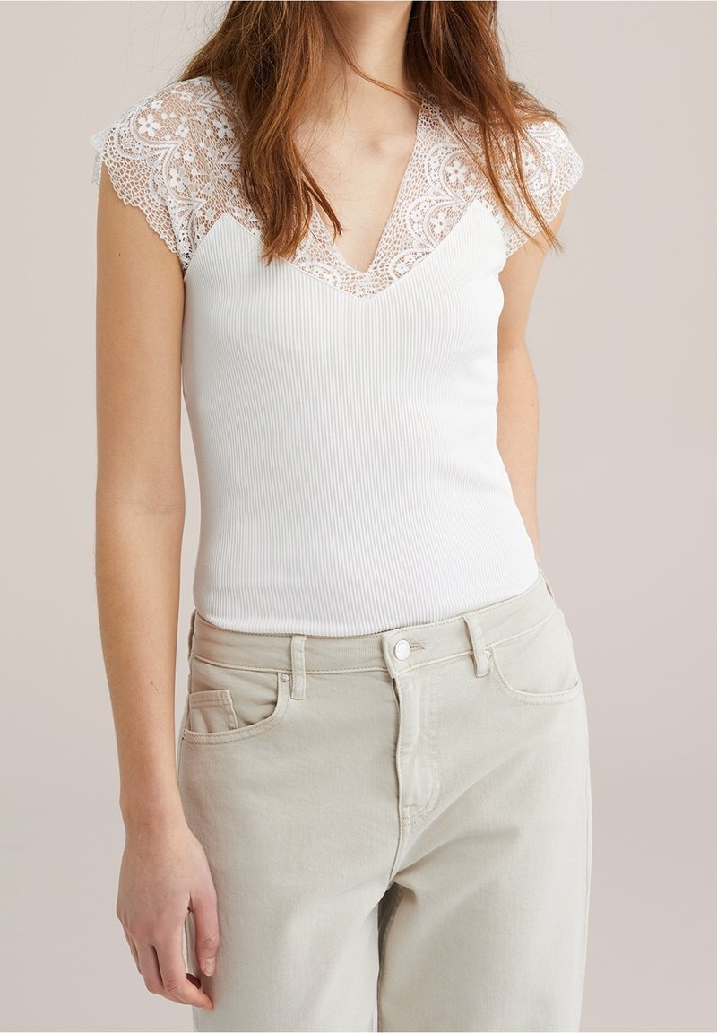 Vit ribbad linne med V-ringning och fina vita spetsärmar, tillsammans med ljusbeige högmidjade byxor.