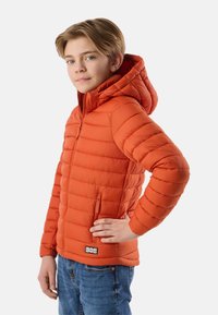 Garçon aux cheveux châtain clair portant une doudoune orange à capuche, debout avec une main sur la hanche et un jean bleu.
