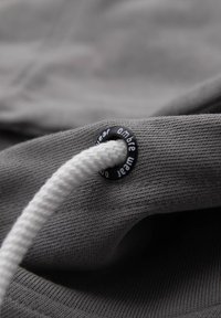 Szara tkanina o teksturowanej powierzchni, z białym sznurkiem przewleczonym przez czarny oczko, z białym napisem, zbliżenie.