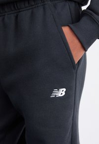 Sorte træningsbukser lavet af blødt stof, med en sidelomme og et broderet hvidt New Balance-logo på underbenet.