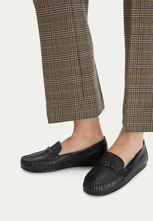 Pies con mocasines de cuero negro combinados con pantalones cortos de cuadros marrones sobre un fondo claro.