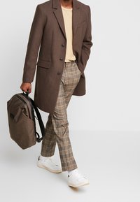Cappotto in misto lana marrone con revers; t-shirt beige chiara; pantaloni a quadri marroni e crema; sneakers bianche; porta uno zaino marrone.