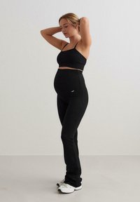 Svart, ribbet graviditet sports-bh og leggings sett, med en tettsittende design og høy midje for komfort. Modellen har på seg hvite treningssko.