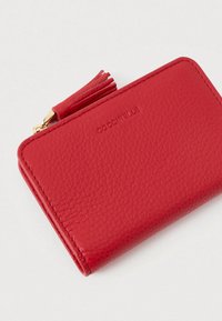 Red textured leather wallet with tassel zipper pull and embossed brand name "Coccinelle" on a white background.