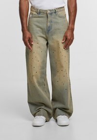 Široke, izblijedele zelenkasto-smeđe traperice s crnim mrljama od boje, nosi se s bijelim tenisicama i bijelom majicom.