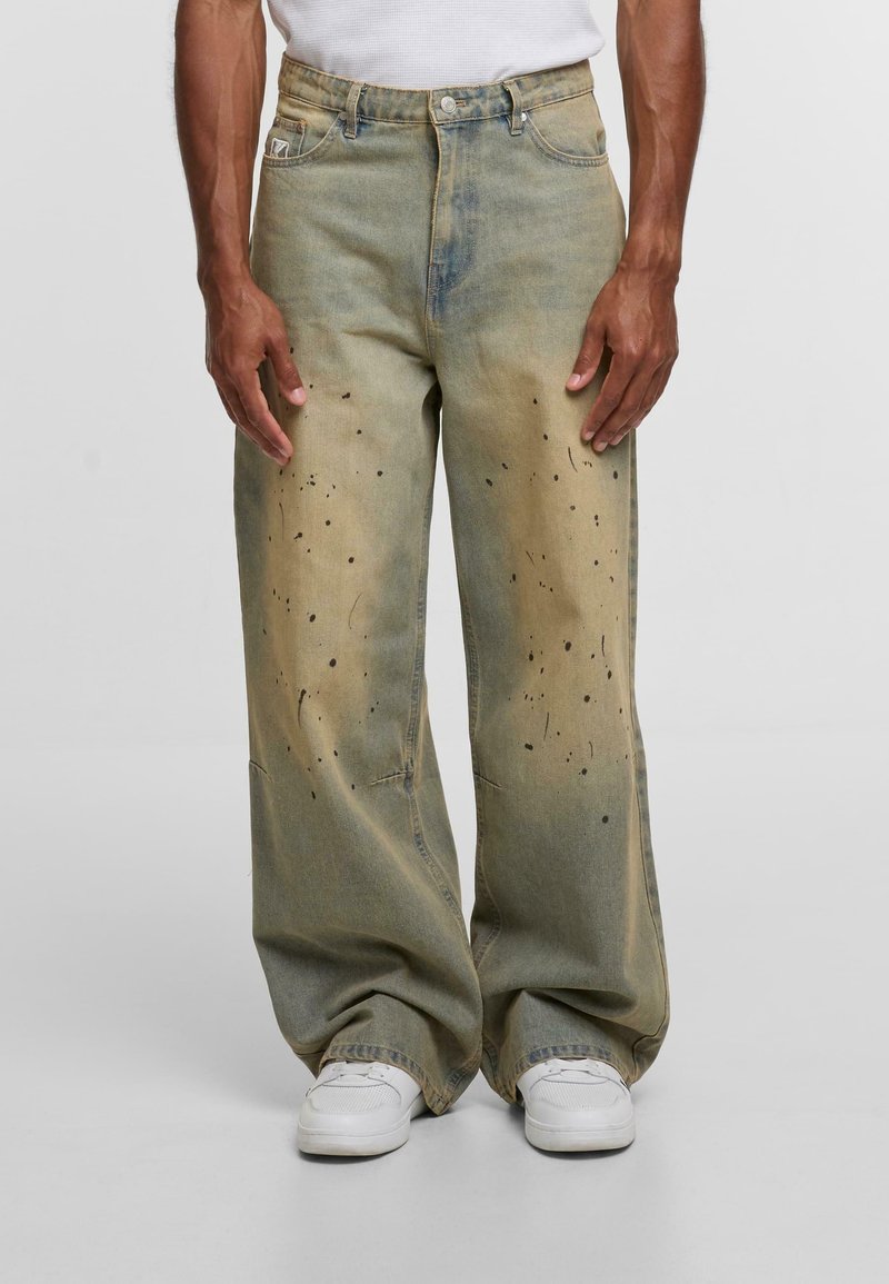 Široke, izblijedele zelenkasto-smeđe traperice s crnim mrljama od boje, nosi se s bijelim tenisicama i bijelom majicom.